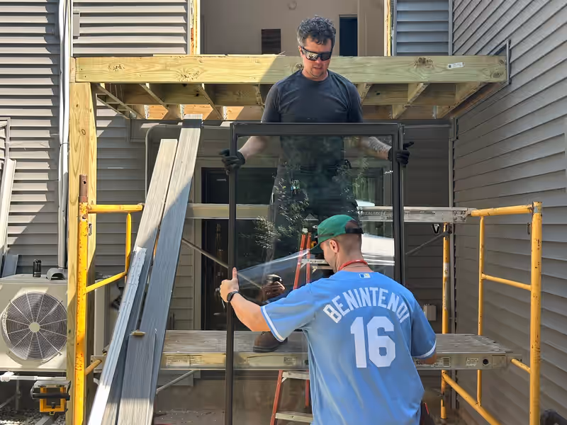 Empirical Renovations crew lifting a glass patio door into a second-story opening above new deck framing in Vermont